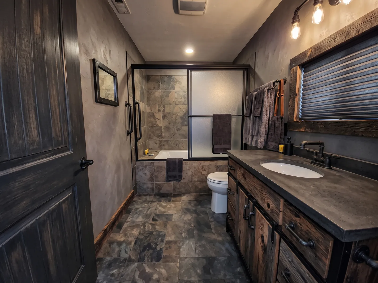 AI generated industrial restyle of the same bathroom: dark plaster walls, dark wood vanity with a stone top, slate floor and exposed-bulb lights. Bath, shower doors, toilet and window stay in place.