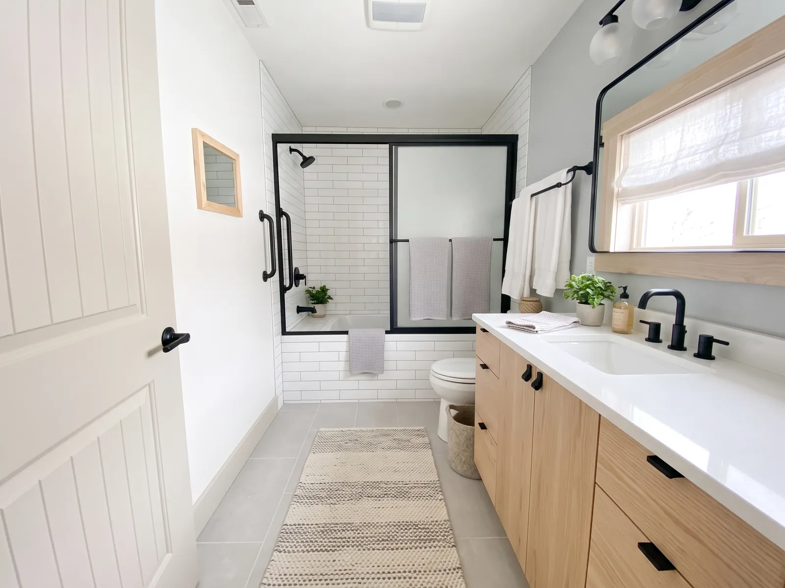 AI generated Scandinavian restyle of the same bathroom: white metro tiles in the shower, white door, light oak vanity, grey floor tiles with a rug and a round black mirror. Bath, shower doors, toilet and window stay in place.