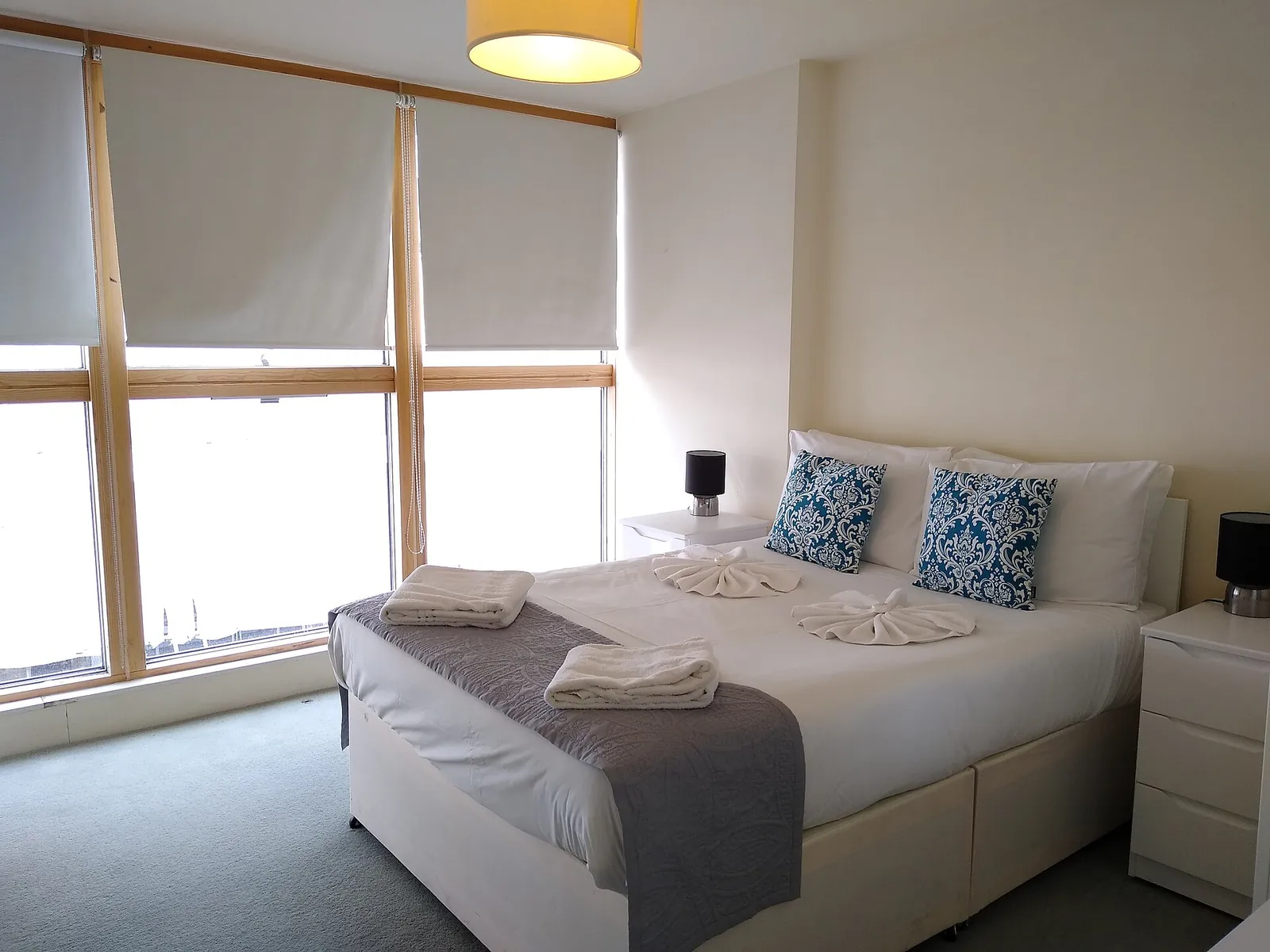 Before: bedroom with a wall of timber-framed windows, white roller blinds and a double bed.
