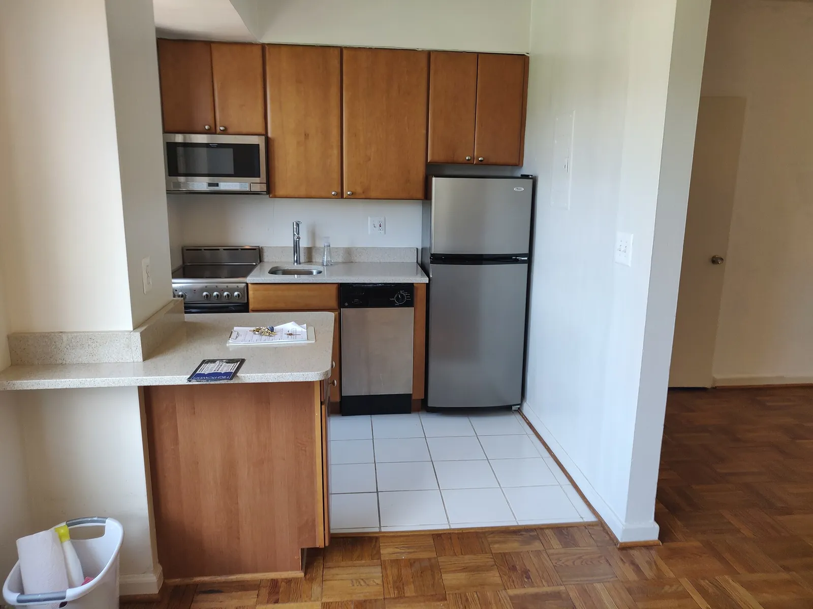 Before: small apartment kitchen with wood cabinets, a steel fridge, white floor tiles and a breakfast bar.