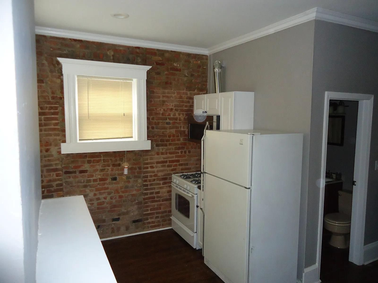 Before: kitchen with an exposed brick wall, a white-framed window, white fridge and gas cooker, and a door to a bathroom.