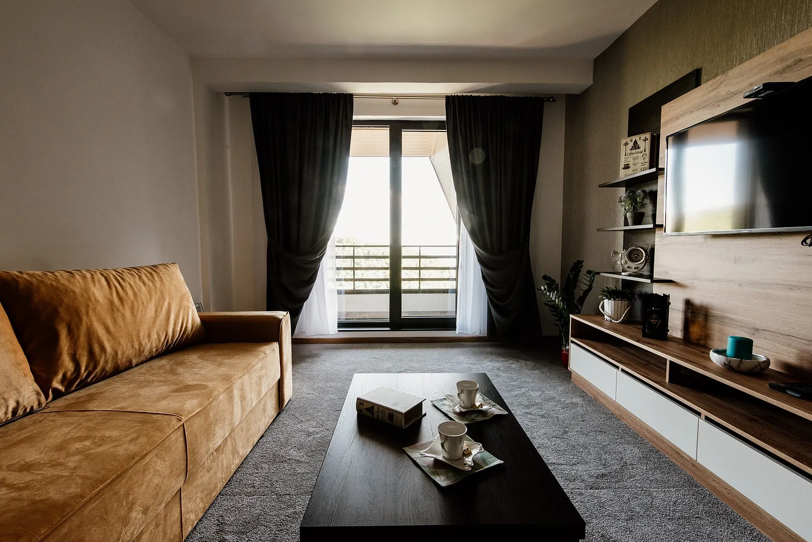 Before: living room with a tan sofa, dark curtains at a balcony door and a wooden TV wall.