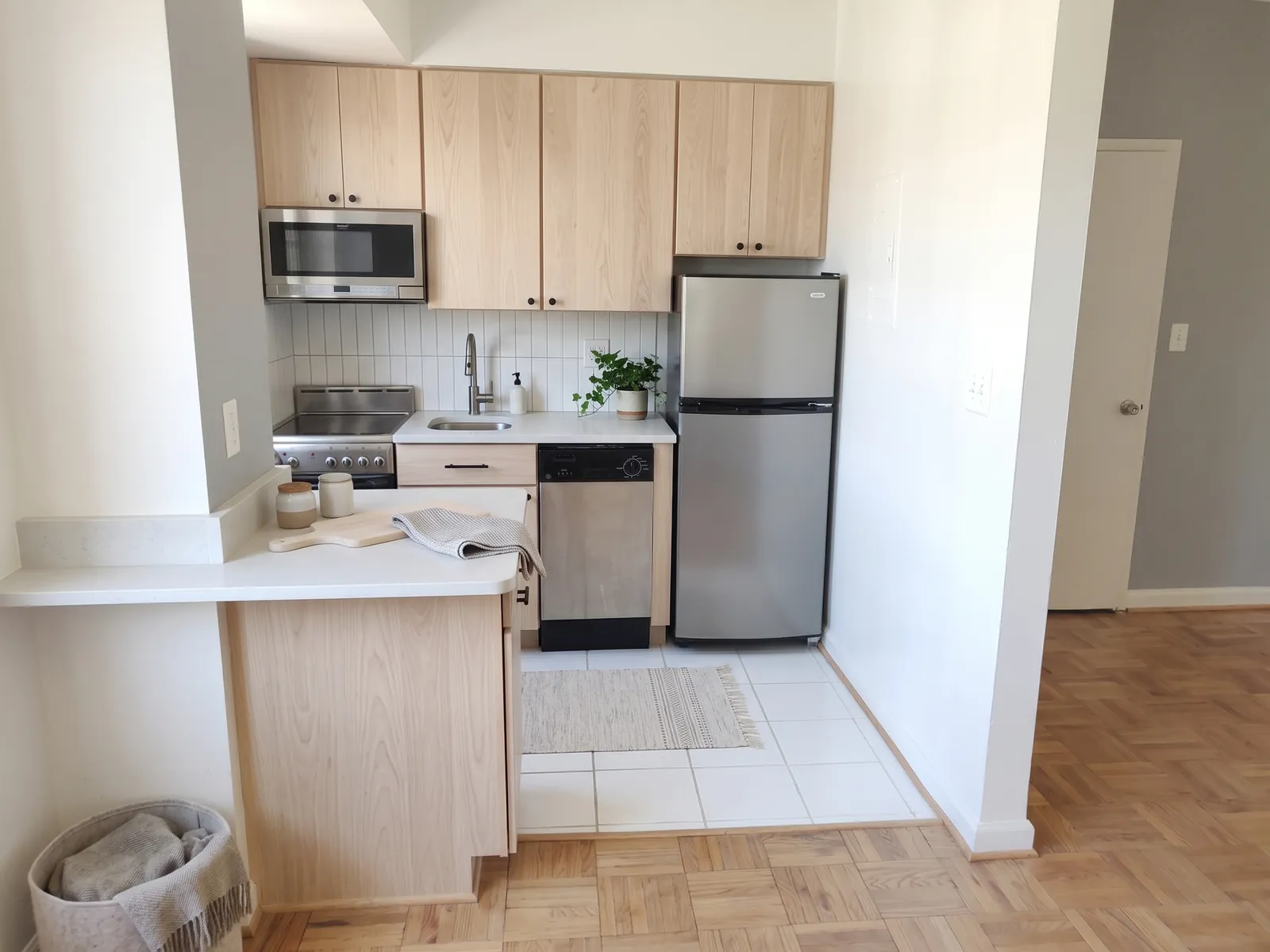 AI generated Scandinavian restyle of the same kitchen: pale oak cabinet fronts, white tile splashback, a pale grey wall and a cream rug. Fridge, cooker, dishwasher and worktop stay where they were.