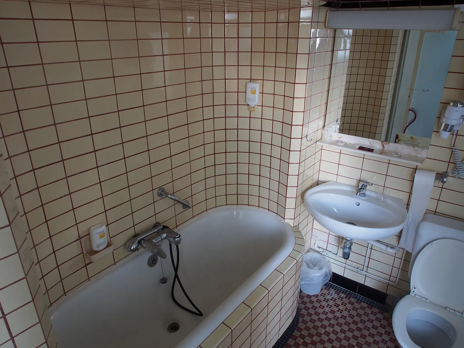 Before: small tiled bathroom photographed with a wide lens, with a curved tiled bath, a wall basin, a mirror and a toilet.